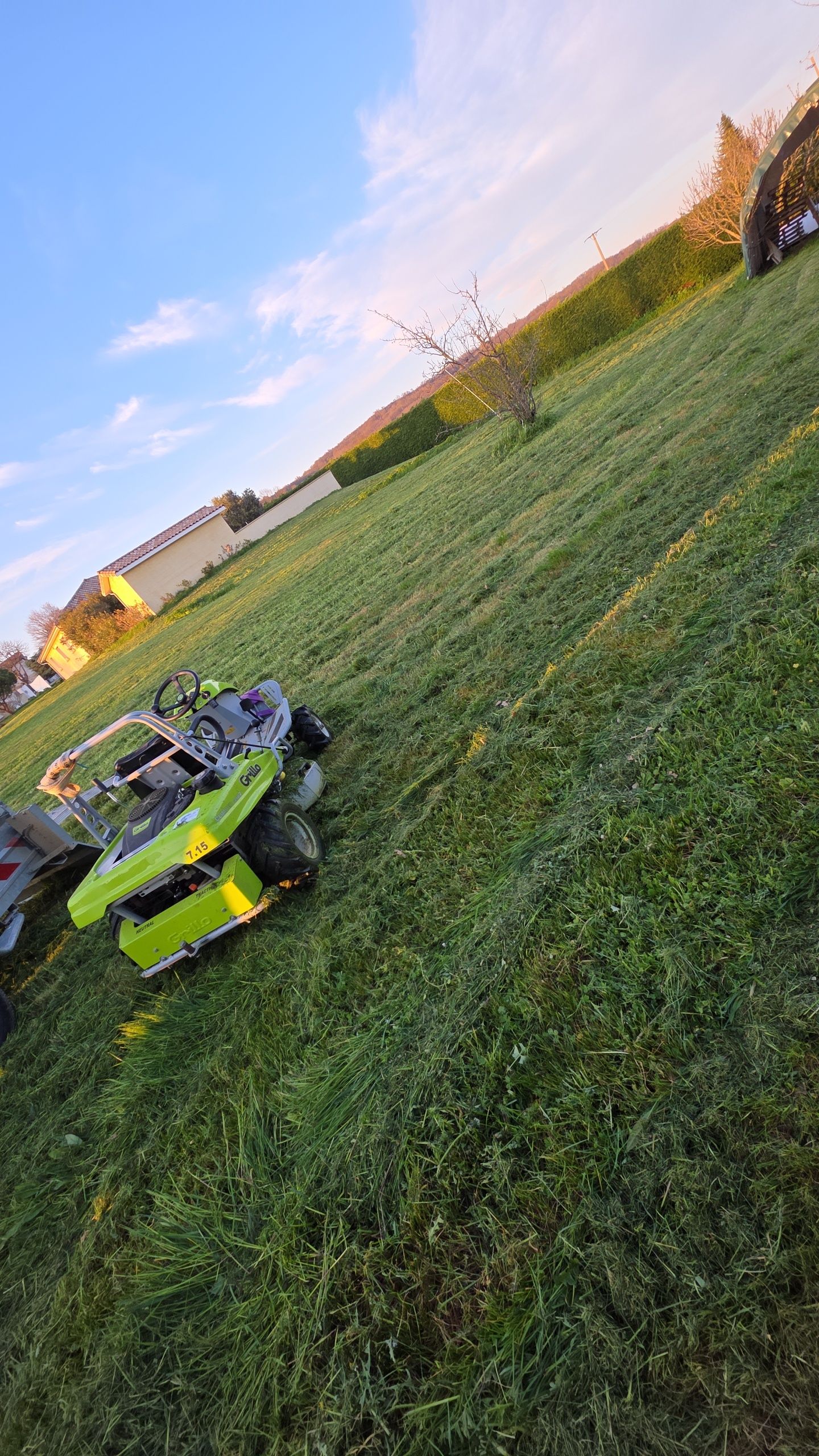 Tondeuse autoportée verte sur un champ herbeux en pente au coucher du soleil, avec des maisons au loin.