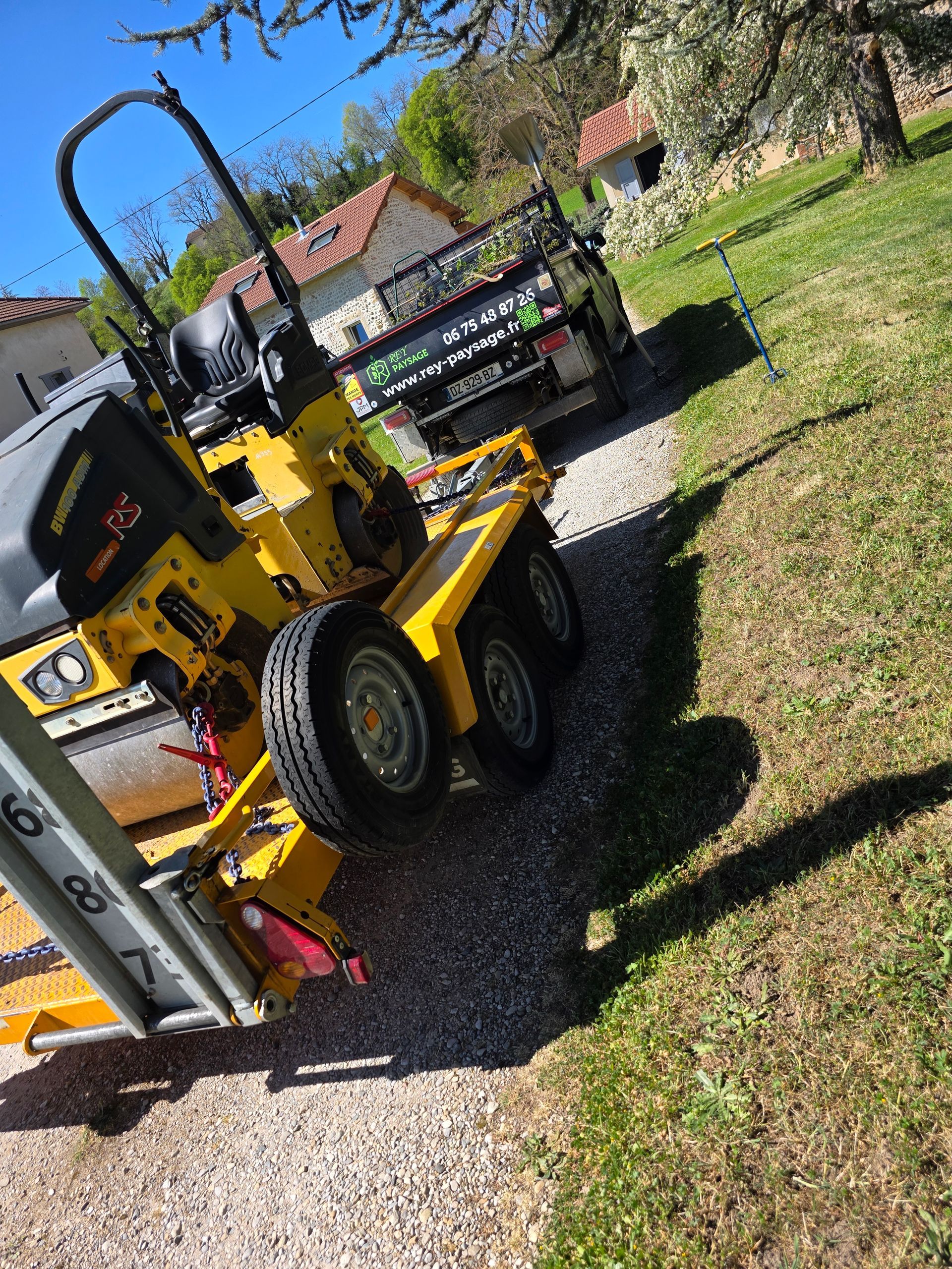 Chargeuse compacte jaune sur une allée de gravier près d'une maison et d'une pelouse par une journée ensoleillée.