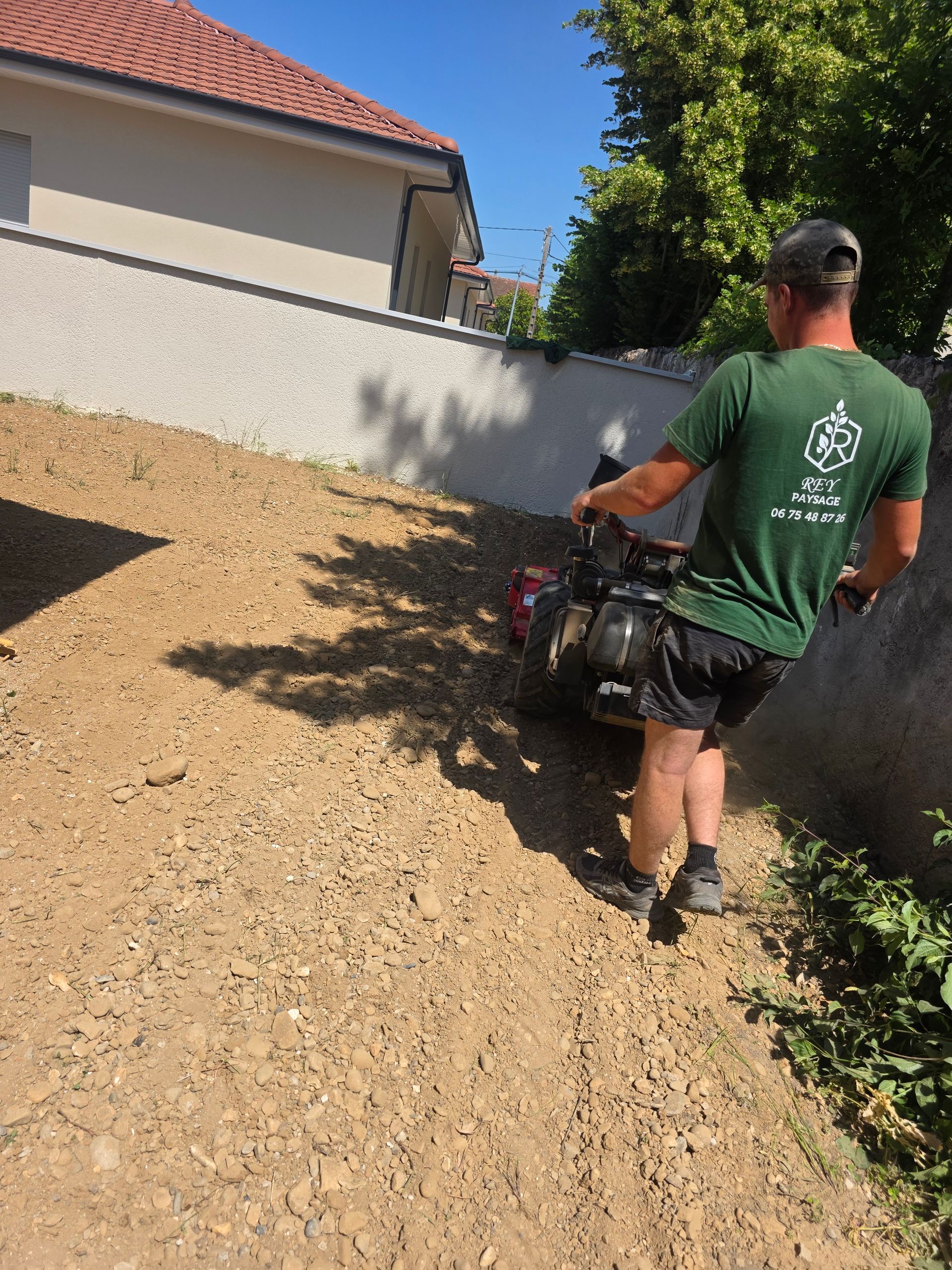 Un ouvrier utilise un outil électrique sur roues sur une allée de gravier longeant une maison et un mur.