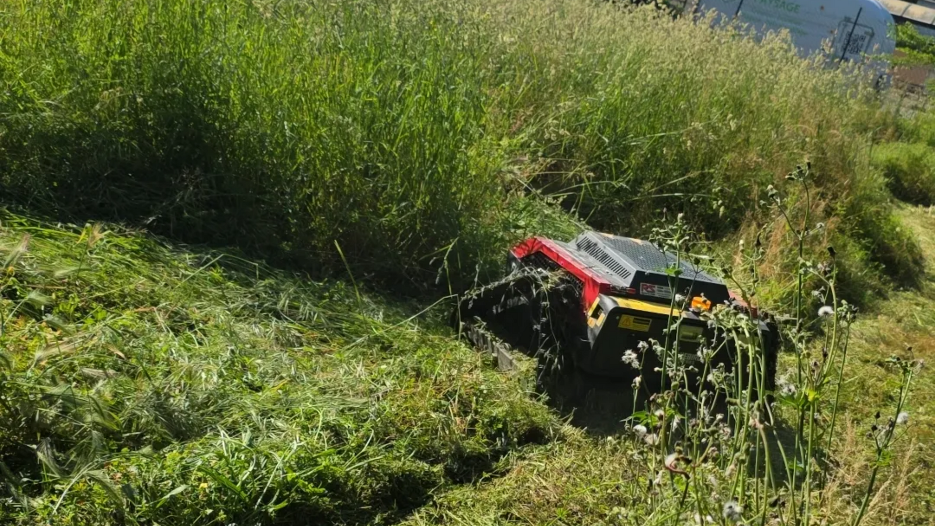 Véhicule tout-terrain rouge et noir partiellement dissimulé dans les hautes herbes vertes d'une colline ensoleillée.