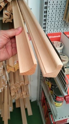 Una mano sostiene dos piezas de moldura de madera clara en el pasillo de una ferretería.