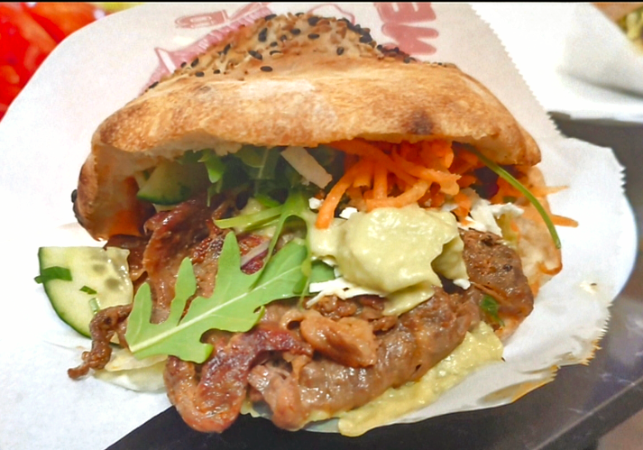 Nahaufnahme eines Kebab-Sandwiches mit Fleisch, Gemüse und Soße auf einem Brötchen, serviert in Papier.