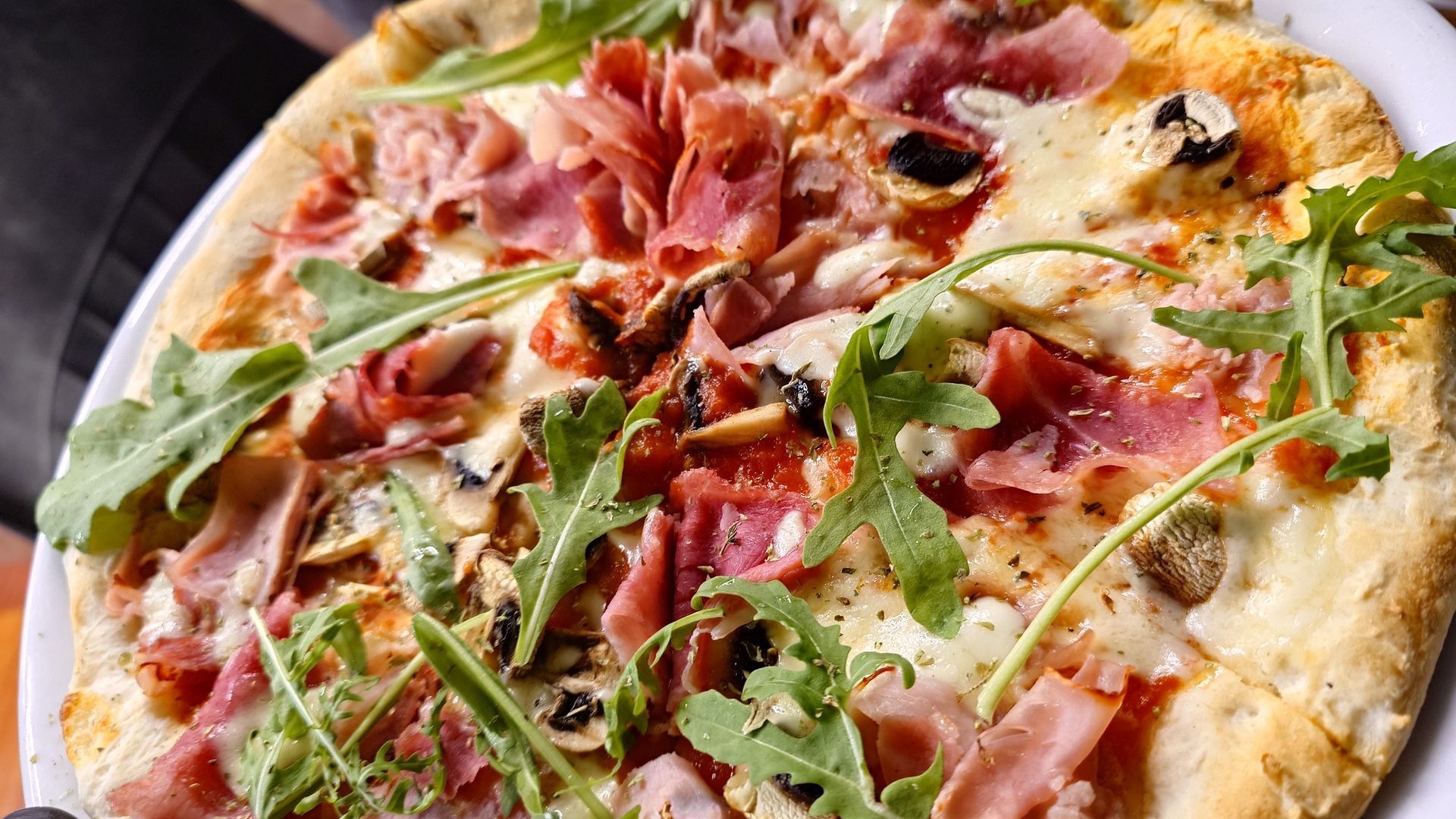 Pizza mit Schinken, Champignons, Käse und Rucola auf einem weißen Teller.