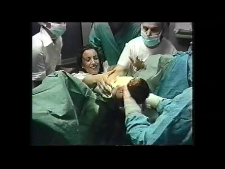 Mujer dando a luz en un hospital, rodeada de personal médico. Sonríe mientras nace el bebé.