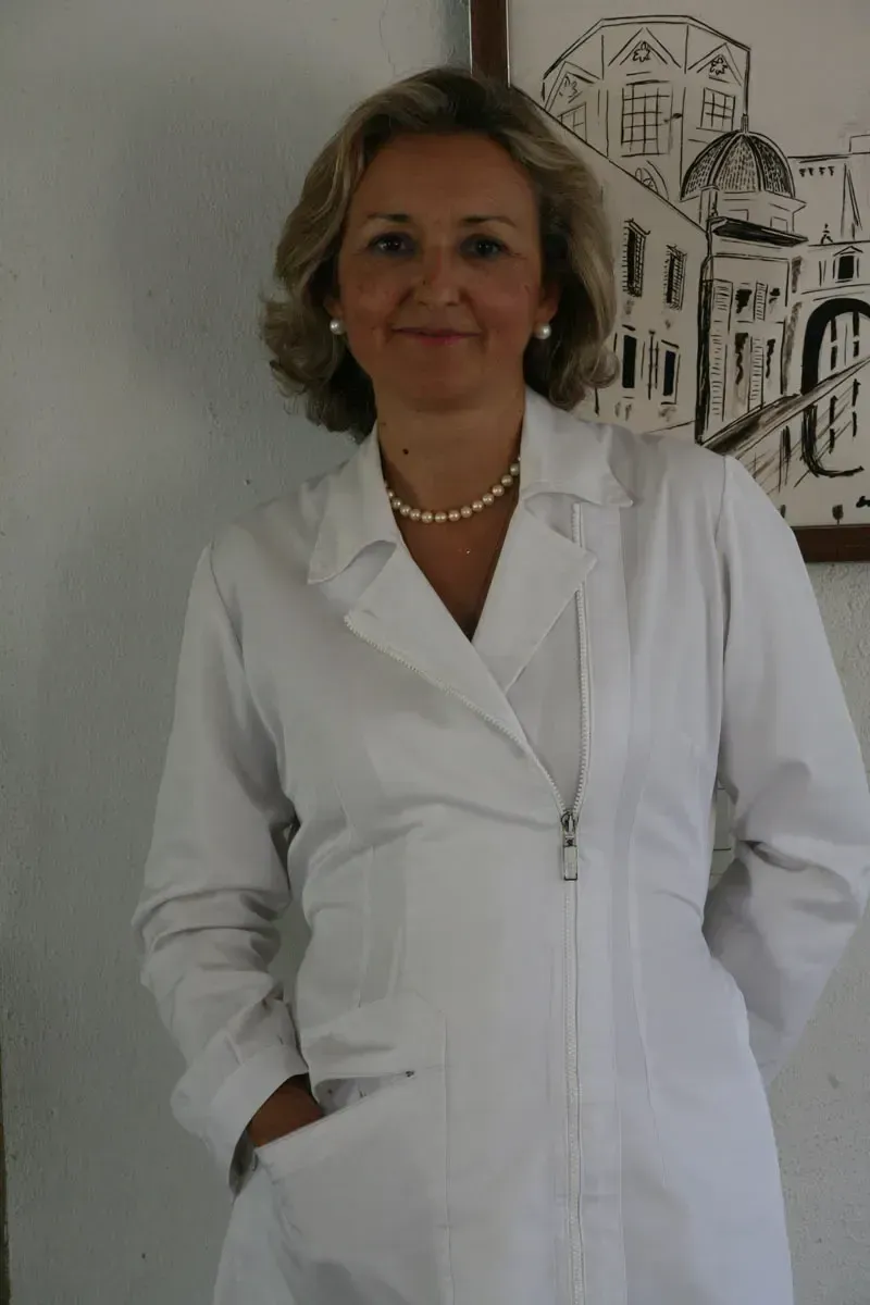 Mujer con bata de laboratorio blanca, sonriendo, con las manos en los bolsillos, de pie en el interior cerca de una obra de arte.