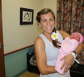 Mujer con un bebé recién nacido, ambos sonriendo. Habitación de hospital. La mujer lleva un vestido blanco y el bebé está envuelto en una manta rosa.