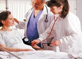 Una enfermera toma la presión arterial a un paciente en la cama mientras un médico observa en un entorno hospitalario.