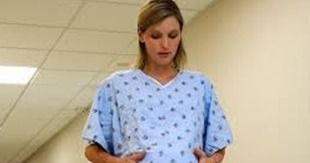Mujer embarazada con bata de hospital, parada en un pasillo, mirando hacia su abdomen.