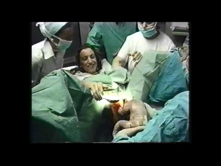 Mujer dando a luz en una habitación de hospital, rodeada de personal médico. Sostienen a su bebé; la mujer sonríe.