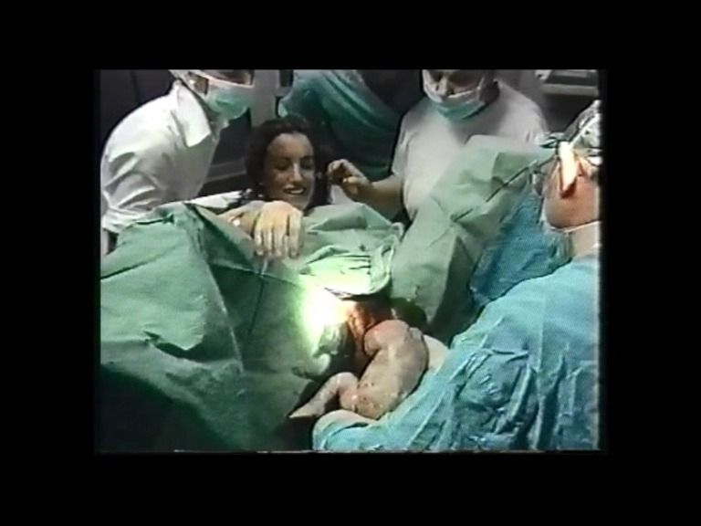 Mujer dando a luz, rodeada de personal médico, sonriendo. Sostienen a su bebé.