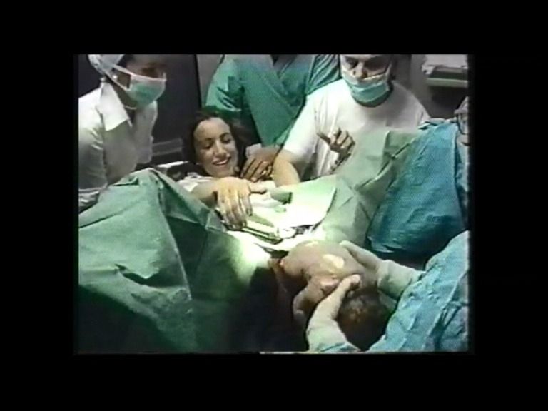 Una mujer da a luz en el hospital, sonriendo. El personal médico la rodea, sosteniendo a un recién nacido.