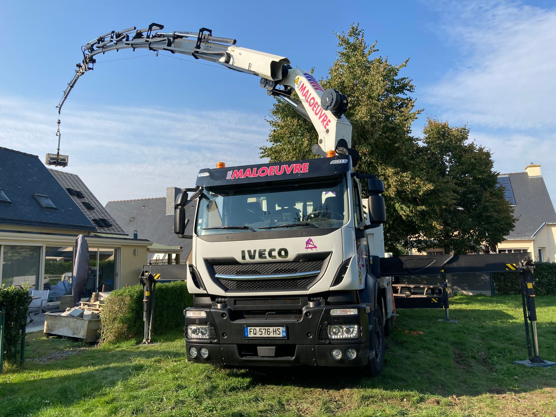 Grue montée sur camion soulevant une charge près d'une maison par une journée ensoleillée.