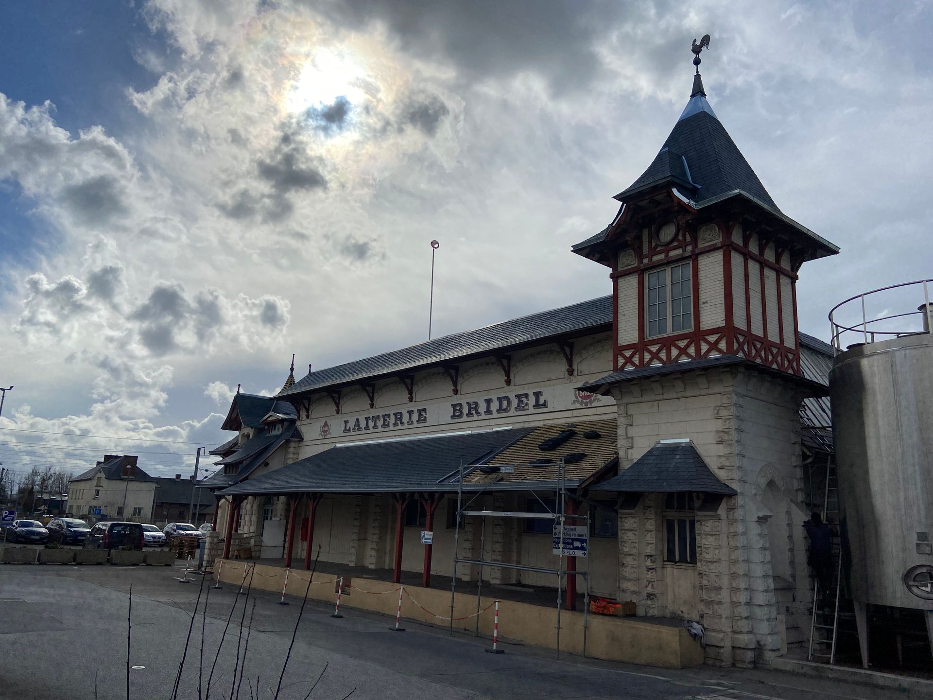 Bâtiment avec tour, portant l'inscription « Laiterie Bridel », sous un ciel nuageux. Parking et réservoirs industriels visibles.