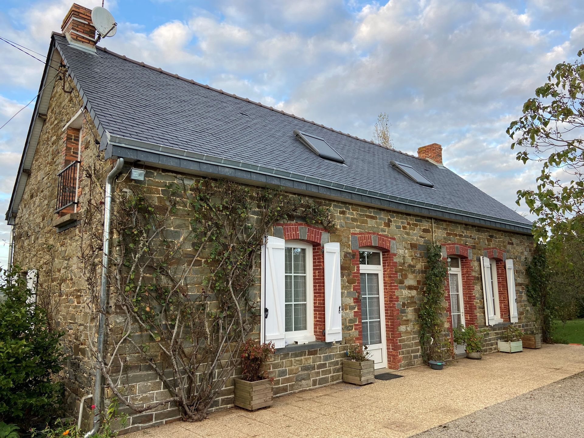 Maisonnette en pierre avec toit en ardoise, volets blancs et allée de gravier.