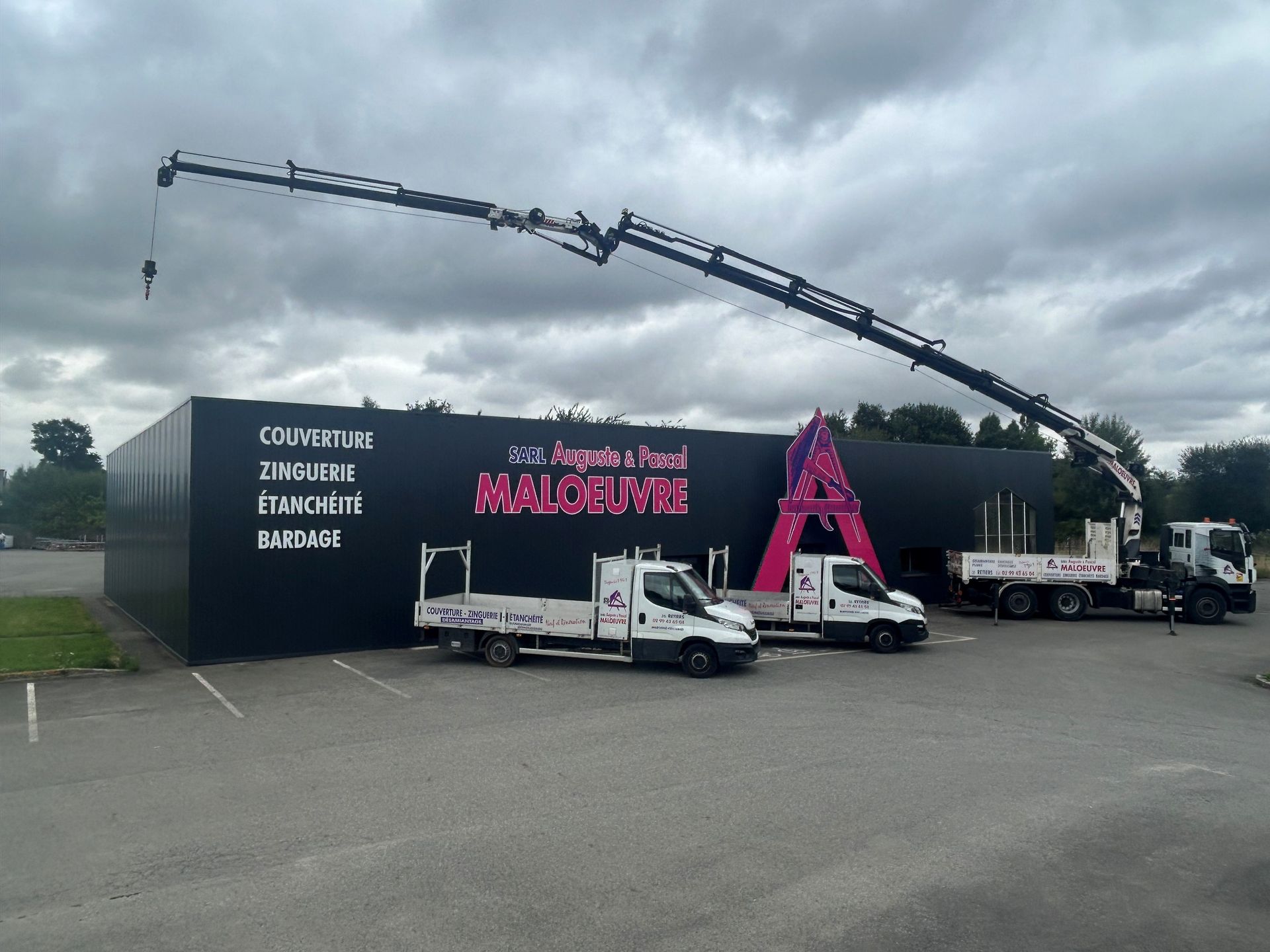 Bâtiment noir avec le logo rose de l'Entreprise Maloeuvre, camions et grue se détachant sur un ciel couvert.