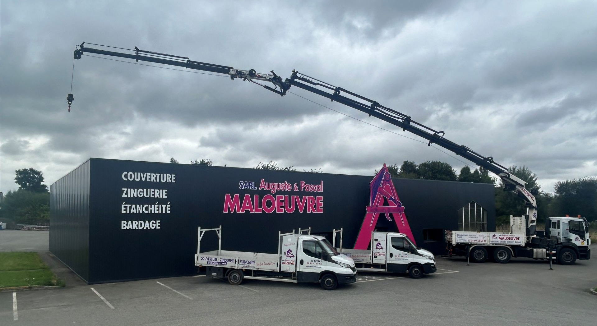 Bâtiment noir avec le logo rose de l'Entreprise Maloeuvre, camions et grue se détachant sur un ciel couvert.