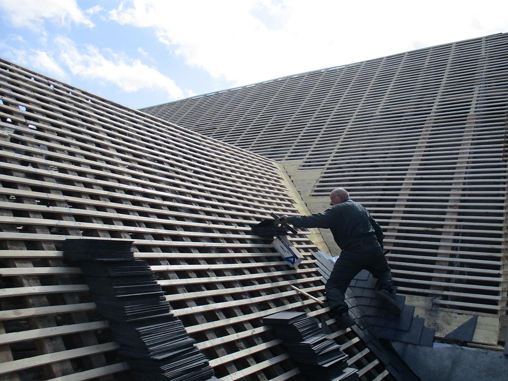 Un homme pose des ardoises sur un toit en pente soutenu par des supports en bois. 
