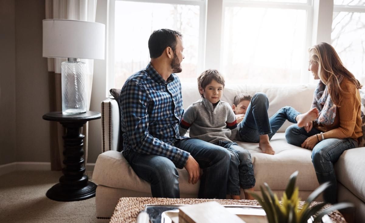 Una familia sentada en un sofá en una luminosa sala de estar, charlando y relajándose junto a la ventana.