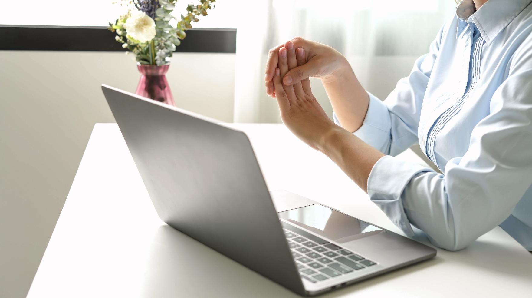 Una persona con una camisa azul claro teclea en una computadora portátil en un escritorio luminoso con flores cerca.