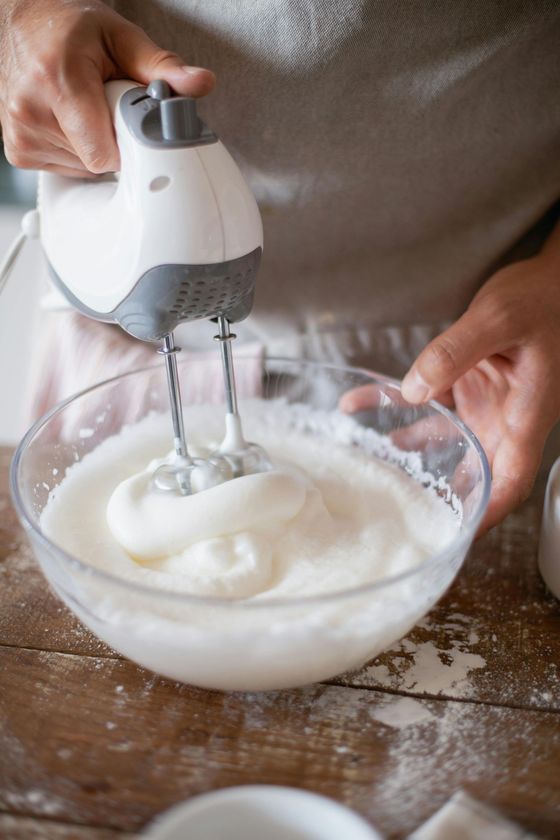 Persona que utiliza una batidora de mano para batir crema en un recipiente de vidrio sobre una superficie de madera.