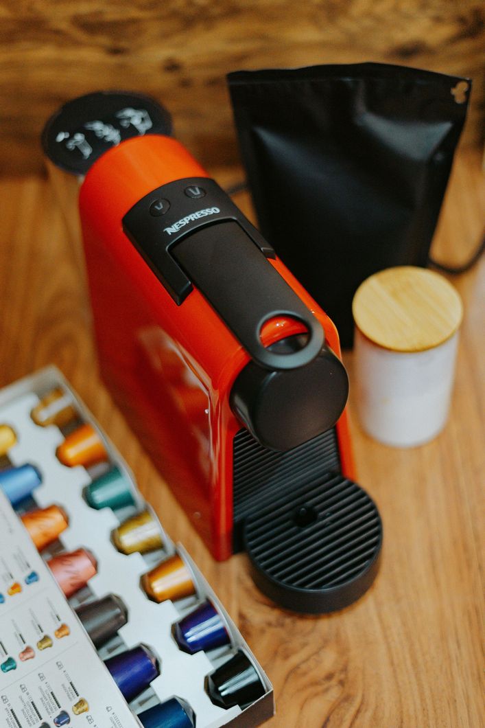 Cafetera Nespresso roja con cápsulas, bolsa negra y bote sobre superficie de madera.