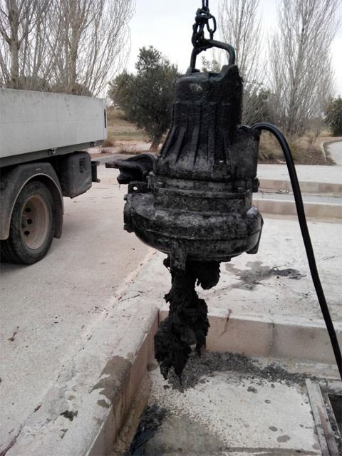 Se está levantando una gran bomba industrial negra de una zanja junto a la carretera, al lado de un camión, con barro en el suelo.