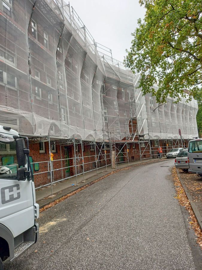 Im Bau befindliches Gebäude, verhüllt mit Gerüsten und weißen Netzen; LKW links, Autos rechts.