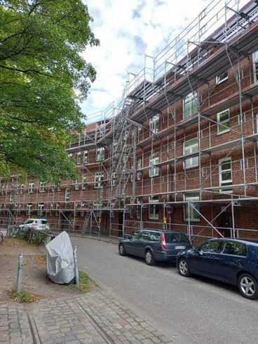 Gebäudefassade mit Renovierungsgerüst; Autos parken auf der Straße bei bedecktem Himmel.