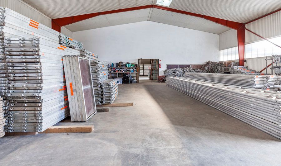 Lagerhalleninnenraum mit gestapelten Metallgerüsten und Ausrüstung; Betonboden, weiße Wände.