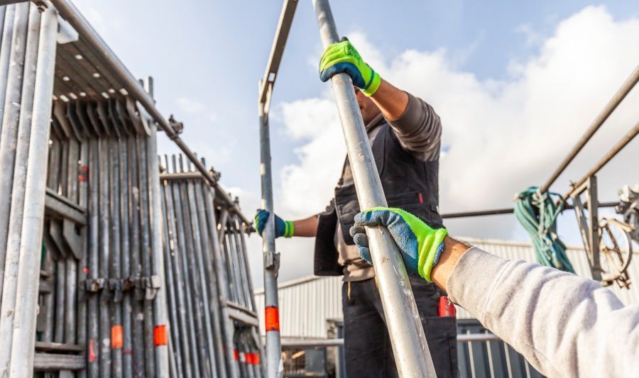 Zwei Arbeiter errichten im Freien ein Gerüst; sie tragen Handschuhe.
