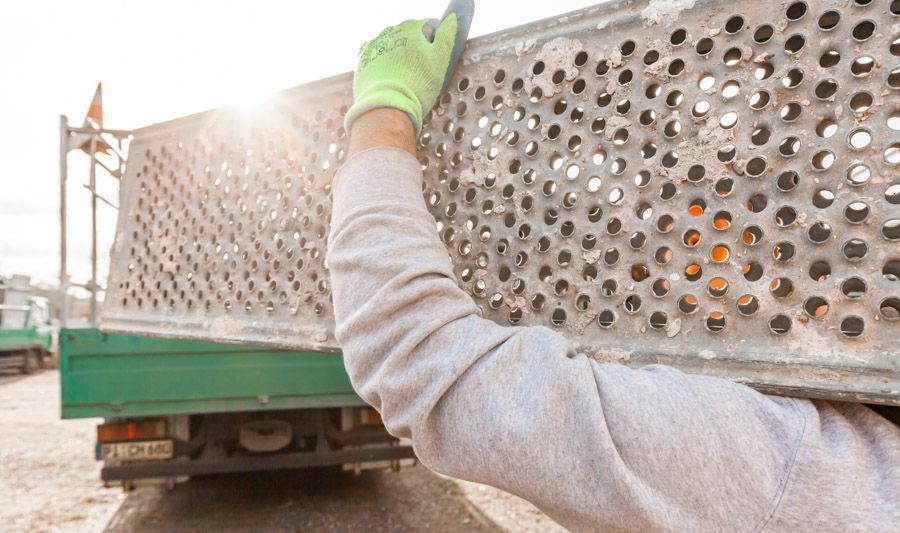 Eine Person hebt im Freien mit einem grünen Handschuh eine perforierte Metallplatte der Ladefläche eines Lastwagens an.