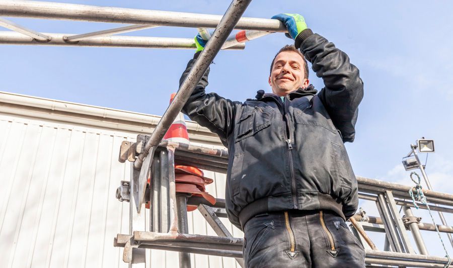 Mann auf einem Gerüst, lächelnd, greift an einem sonnigen Tag nach einer Metallstange.