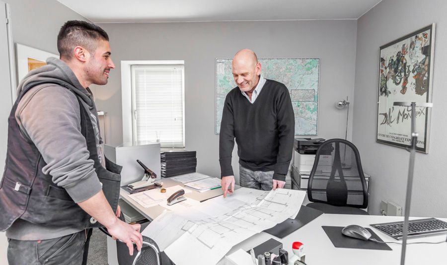 Zwei Männer sitzen in einem Büro und studieren Baupläne an einem Schreibtisch. Der eine lächelt, der andere schaut zu. Graue Wände, Kunstwerke.