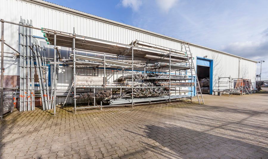 Außenansicht eines Lagerhauses mit Gerüstregalen. Blaue Tür geöffnet. Graue Metallverkleidung, gepflasterter Parkplatz.