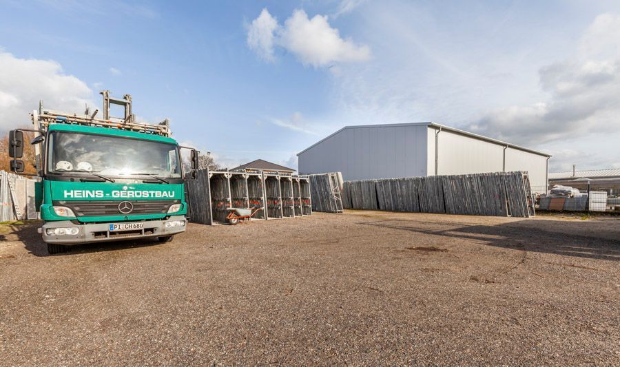Ein grüner Lastwagen parkt auf einem Schotterplatz zwischen verschiedenen Betonbauten und einer großen Lagerhalle unter blauem Himmel.