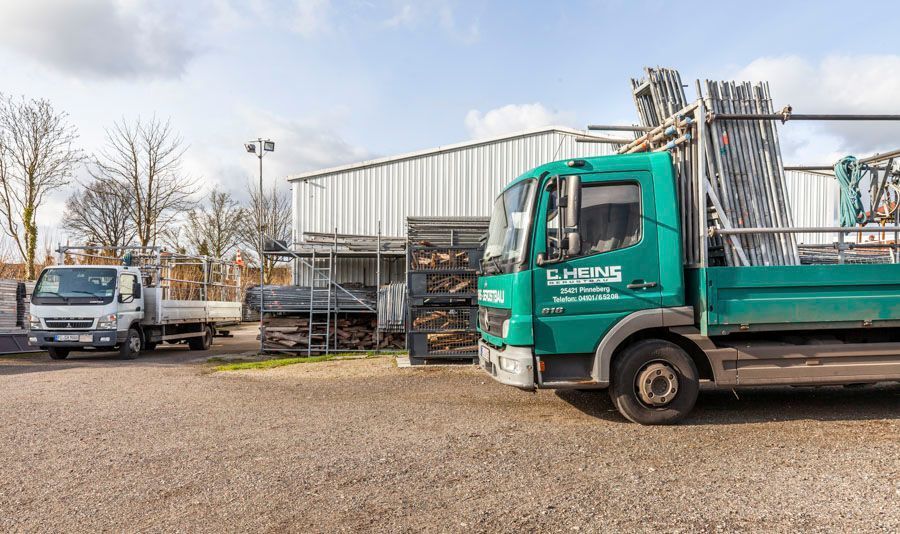 Zwei Lastwagen auf einem Hof mit einem Gebäude; ein grüner Lastwagen, beladen mit Metallgerüsten.