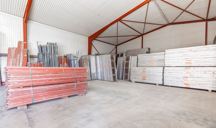 Innenansicht einer Lagerhalle mit gestapelten Baumaterialien; Betonboden, Metallträger und Wellblechdecke.