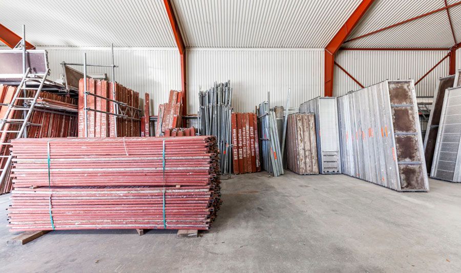 Innenansicht einer Lagerhalle mit gestapelten roten Metallträgern und verschiedenen Baumaterialien.