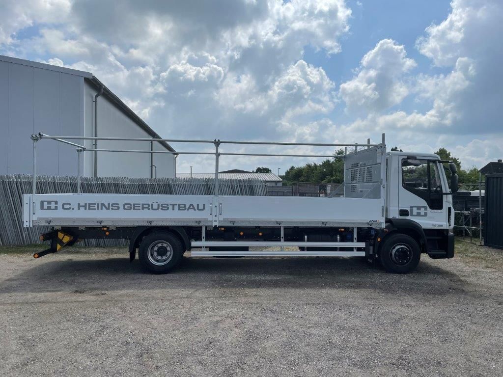 Weißer Pritschenwagen mit Gerüst, geparkt vor einem Gebäude, sonniger Himmel.