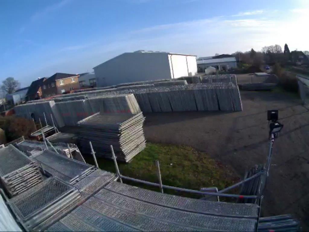 Ein Stapel Metallgerüstplatten auf einem Industriegelände, im Hintergrund Gebäude und ein klarer Himmel.