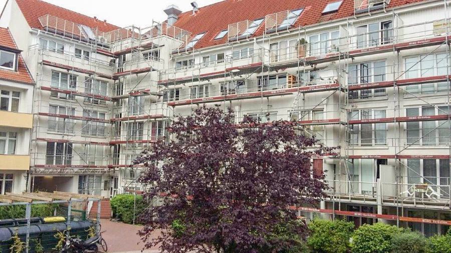 Die Fassade eines Gebäudes wird renoviert; im Vordergrund sind ein Gerüst, ein rotes Dach, Fenster und ein purpurblättriger Baum zu sehen.