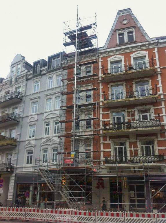 Gerüst an einem Backsteingebäude wegen Reparaturarbeiten; rot, weiß, grau und braun. Ladenfronten im Erdgeschoss, Straßenabsperrung davor.