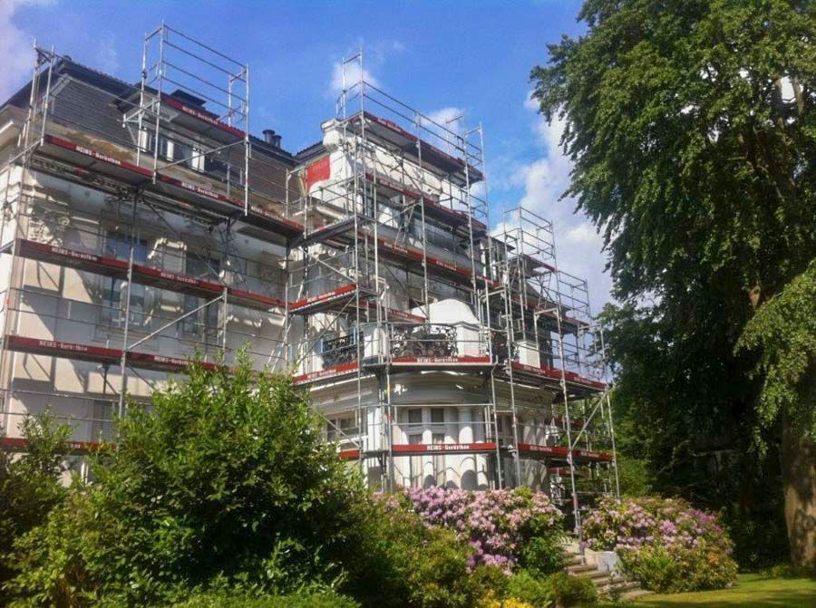 Ein mehrstöckiges Gebäude, das derzeit renoviert wird, ist von einem Gerüst umgeben; man sieht es von einem Garten mit Bäumen und Sträuchern aus.