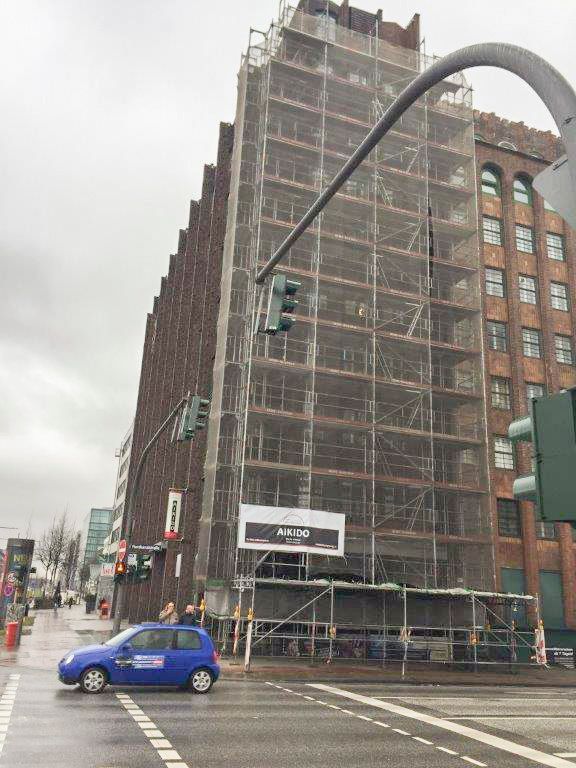 Ein hohes Backsteingebäude ist eingerüstet, ein blaues Auto überquert die Straße. Bewölkter Tag.