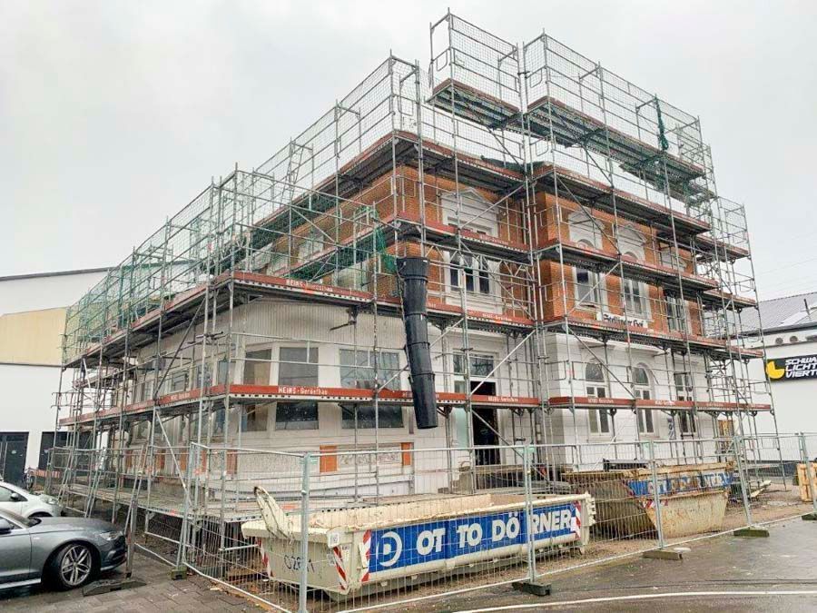 Ein im Bau befindliches Gebäude mit Gerüst und Netzen. Fassade aus Ziegeln und weißen Elementen. Davor steht ein Müllcontainer.