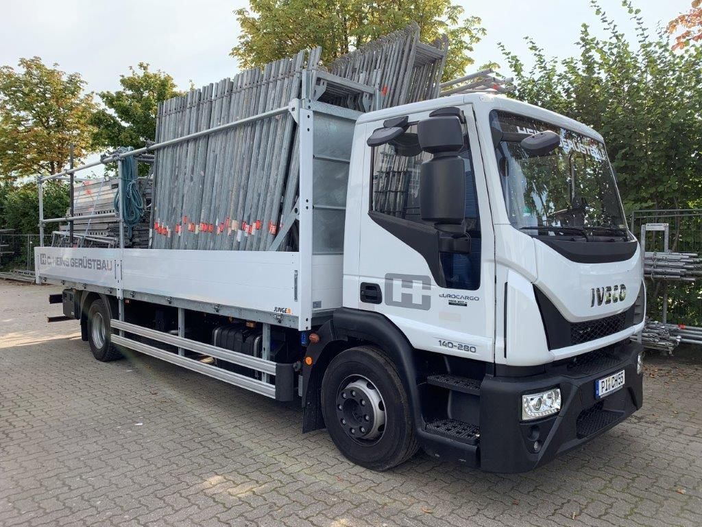 Ein weißer Iveco-Lkw, beladen mit Gerüstteilen, steht auf einem gepflasterten Parkplatz.