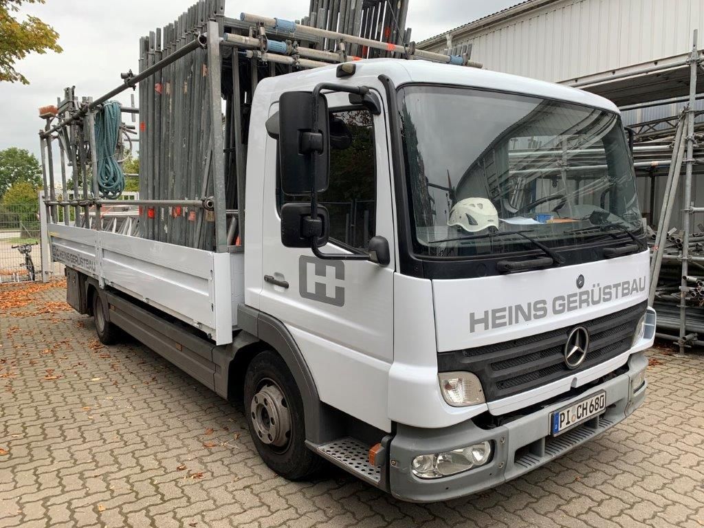 Weißer LKW mit dem Logo „HEINS GERÜSTBAU“, der ein Gerüst auf einer gepflasterten Fläche transportiert.
