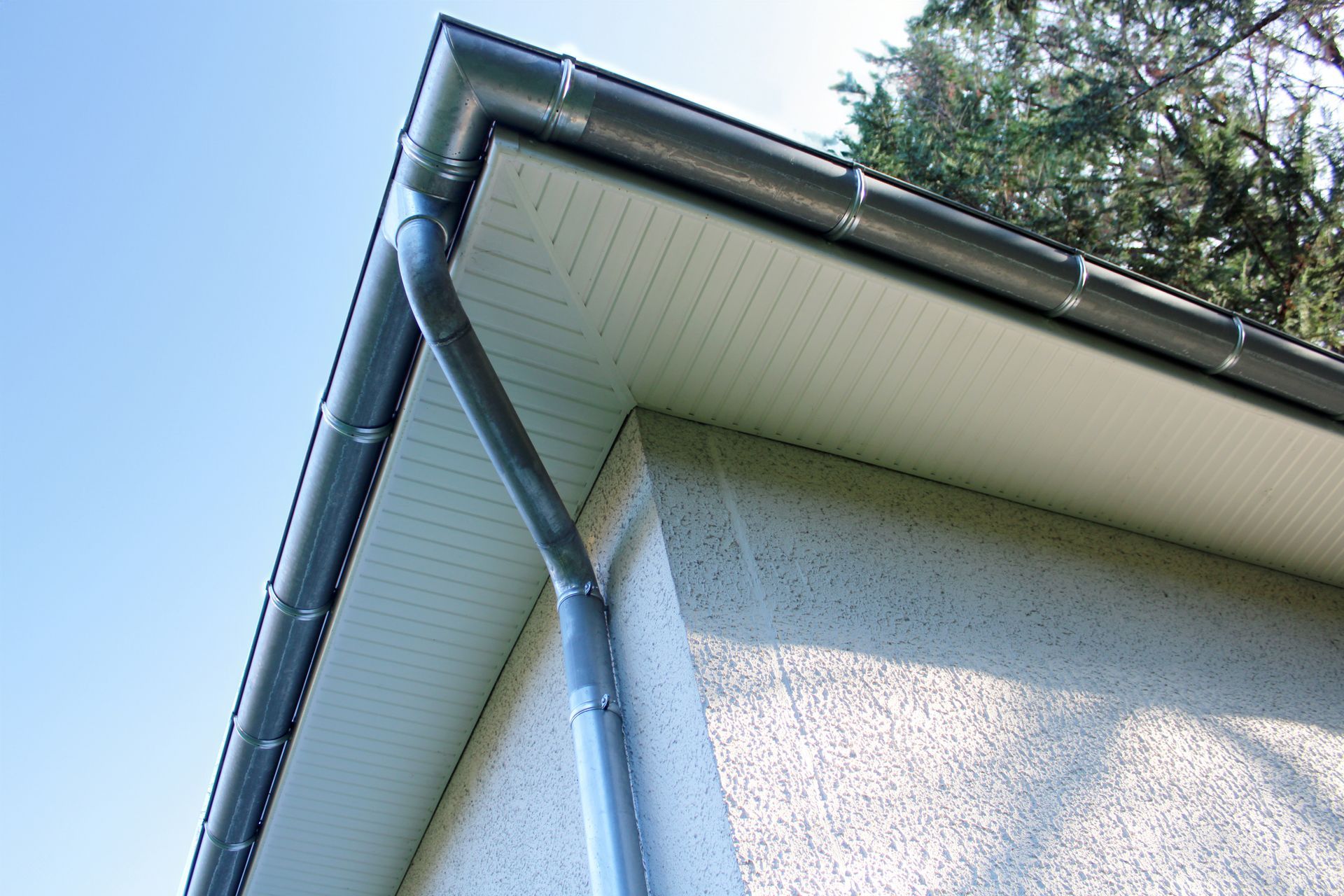 Gouttières et descente pluviale grises sur un bâtiment en stuc blanc, avec un ciel bleu clair en arrière-plan.