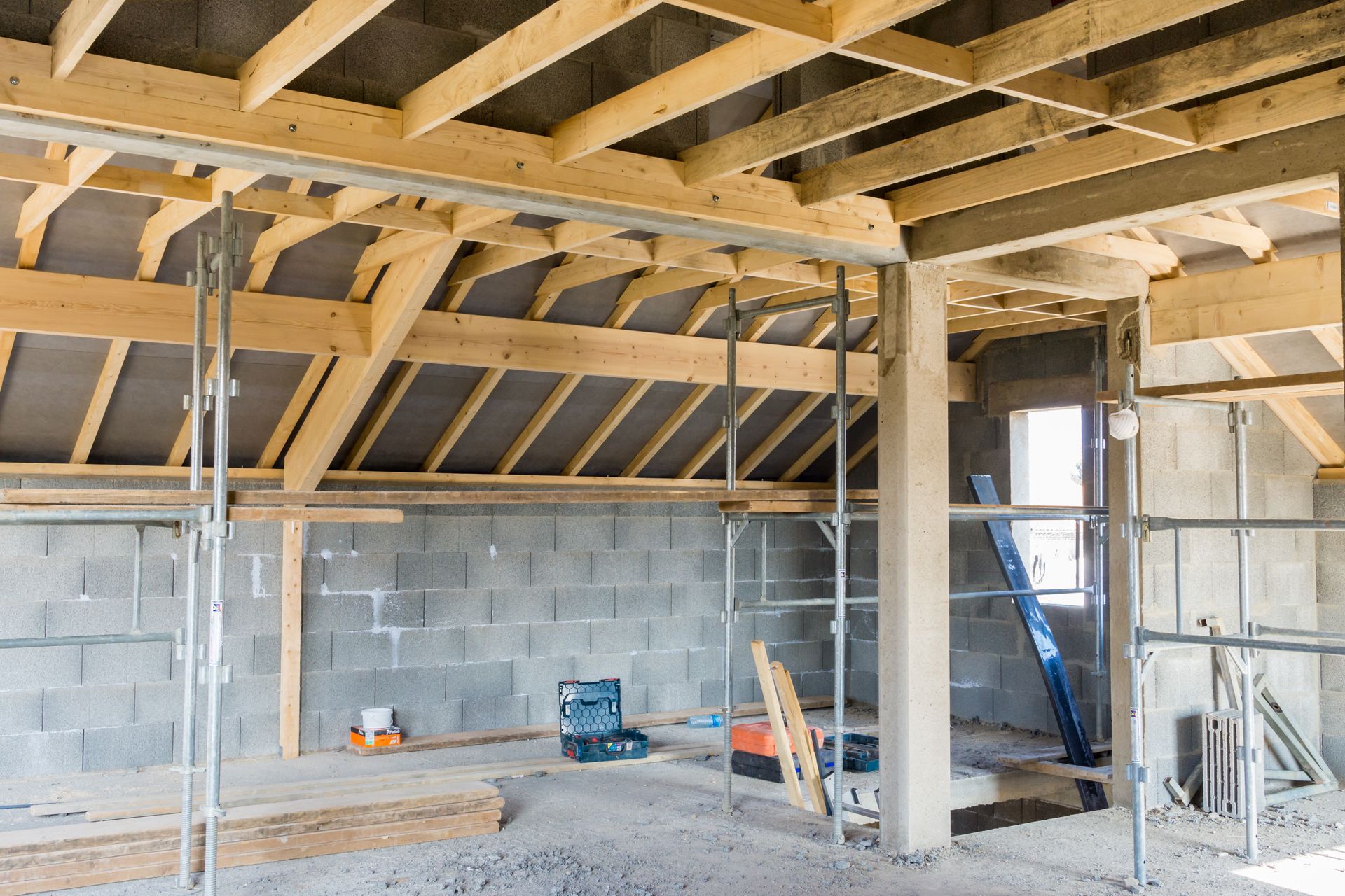 Intérieur d'un bâtiment en construction, montrant les poutres de toit en bois, les murs en parpaings gris et les échafaudages.