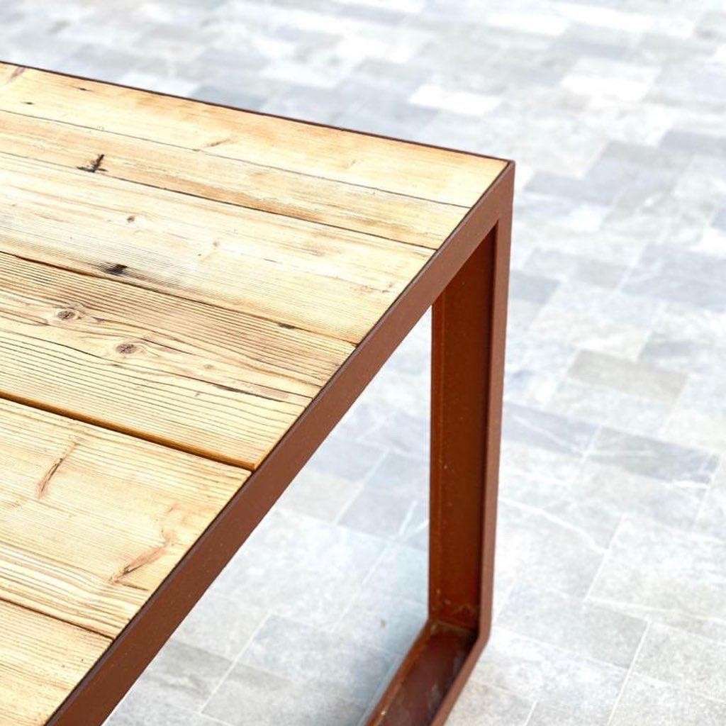 Una mesa de madera rústica con un marco de metal marrón desgastado sobre un fondo de baldosas de piedra gris claro.